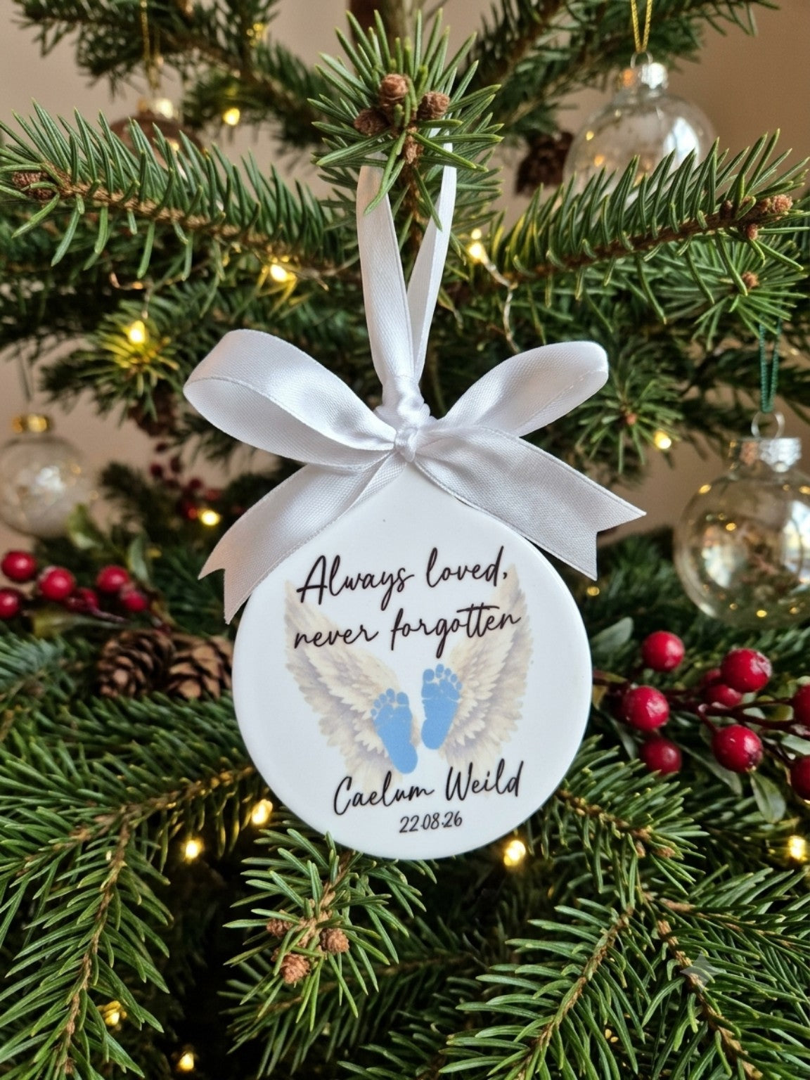 Personalised white memorial bauble hanging on a Christmas tree with angel wings and blue footprints, reading Always loved, never forgotten